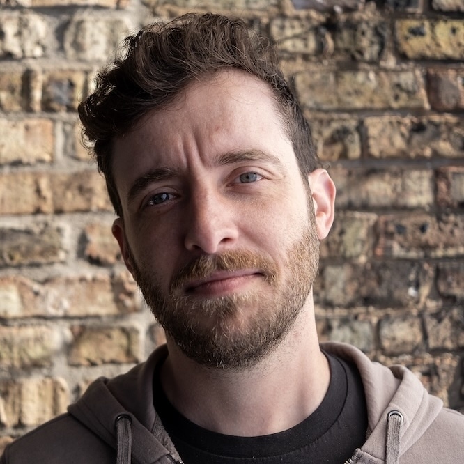 Joshua Bartz standing in front of a worn brick wall and looking at the viewer suspiciously