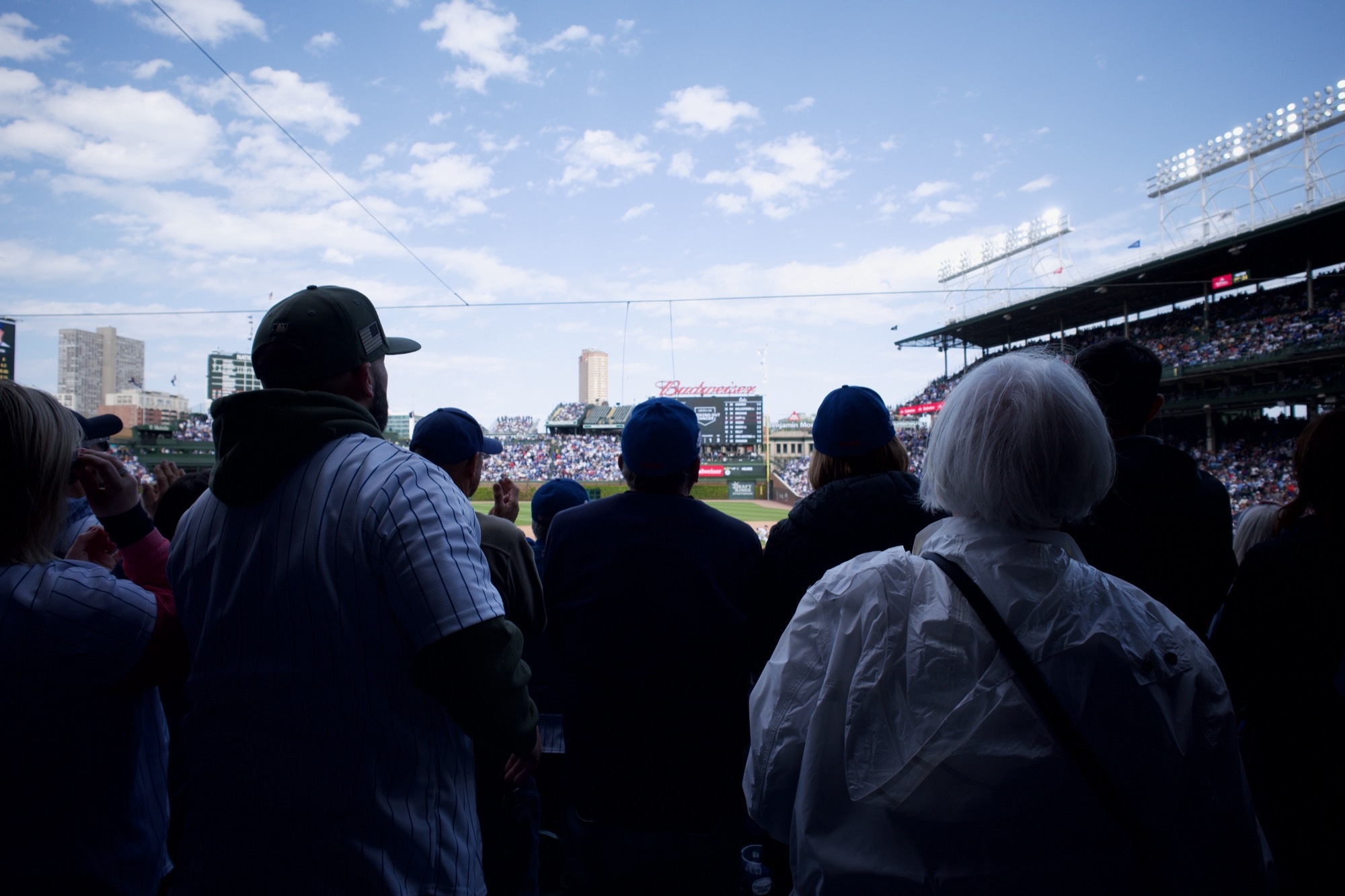 Behind a wall of standing Cubs fans looking on as a play is unfolding on the baseball field ahead.